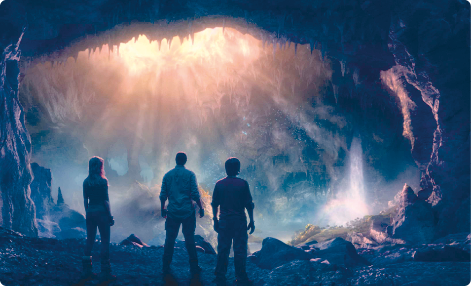 Fotografia. À frente, três pessoas em pé e de costas. Elas estão em um local com muitas rochas ao redor e em cima; em frente a eles, uma cachoeira e uma grande abertura na parte de cima, por onde entram raios de luz.