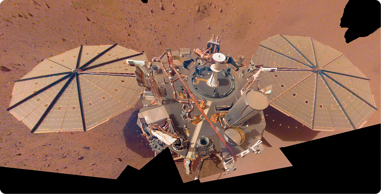 Fotografia. Sonda sobre superfície terrosa. A sonda contém dois discos nas laterais e, no centro, estruturas cilíndricas acopladas em uma base redonda.