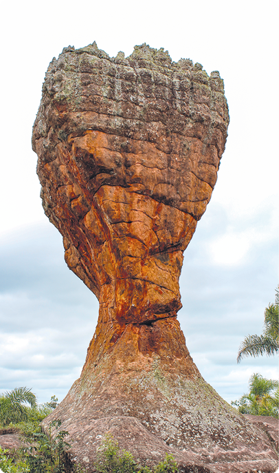 Fotografia deuma grande rocha vertical e amarronzada, com base larga, centro estreito e parte superior mais larga que a base.