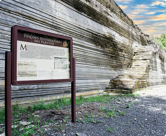 Fotografia. Ao fundo, grande parede de rocha composta por camadas escuras e claras. À frente, uma placa com informações verbais. 