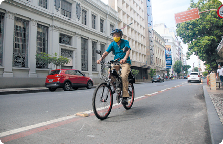 Fotografia. andando de bicicleta, usando capacete e máscara, cobrindo nariz e boca. está rua onde há carros estacionados e construções.