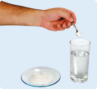 Fotografia. Uma mão segurando uma colher com sal em cima de um copo transparente com água. Ao lado, um pequeno prato com sal.