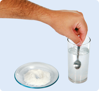 Fotografia. Uma mão segurando uma colher dentro de um copo transparente com água. Ao lado, um pequeno prato com sal.