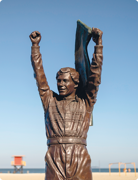 Fotografia. Uma estátua marrom de um homem com os braços erguidos, usando um macacão de mangas compridas e segurando uma bandeira com o braço esquerdo.