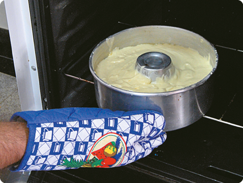 Fotografia. Mão de uma pessoa, usando luva térmica, colocando uma forma de alumínio com massa amarelada de líquido denso dentro do forno de um fogão. 