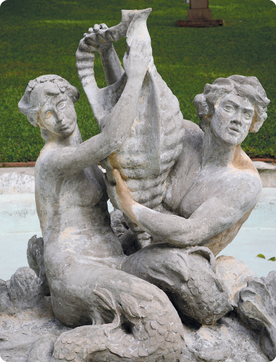 Fotografia. Uma estátua com duas pessoas com cauda de peixe, segurando um vaso grande. As estátuas são acinzentadas e com manchas amarronzadas.