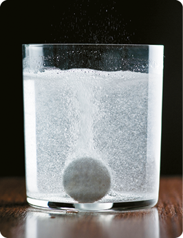Fotografia. Um copo transparente com água e um comprimido efervescente que causa bolhas.