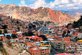 Fotografia. Vista aérea de uma cidade com muitas casas e prédios de várias cores. Ao fundo e à esquerda inferior, montanhas com pouca vegetação.