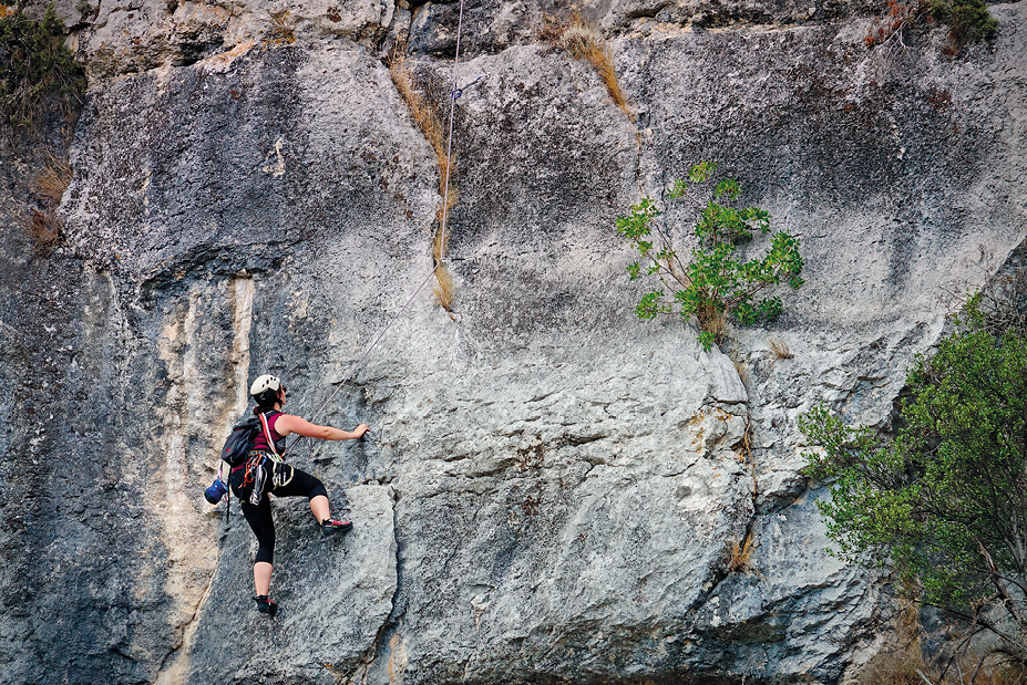 Fotografia. Uma pessoa com capacete, cordas presas ao corpo e mochila nas costas, escalando uma enorme pedra acinzentada e com vegetação.