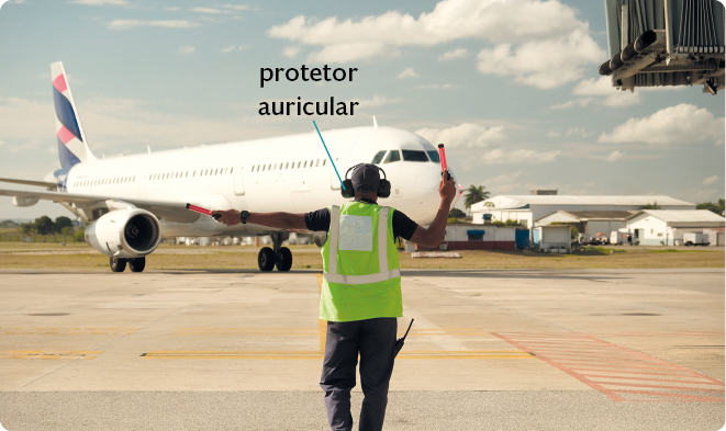 Fotografia. Um homem no aeroporto na frente de um avião. Ele usa um protetor auricular, um colete verde e segura um pequeno bastão laranja com o braço direito erguido, enquanto segura outro bastão laranja com o braço esquerdo esticado. Ele também carrega um rádio comunicador em sua calça.