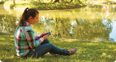 Fotografia. Uma mulher sentada em um gramado de frente para um lago. Ela segura, com as duas mãos, um aparelho eletrônico; uma perna está dobrada e a outra esticada, sua cabeça está inclinada para frente, olhando o aparelho.