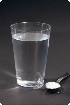 Fotografia. Um copo transparente com água e, ao lado, uma colher com sal.