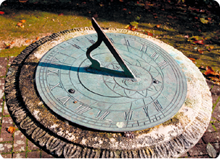 Fotografia. Relógio de sol com base circular achatada e azulada sobre uma pedra. No centro, ponteiro para cima, fazendo sombra sobre a base, à esquerda.