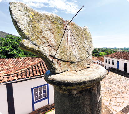 Fotografia. À frente, uma escultura de pedra de um relógio de sol. Estrutura achatada sobre uma base cilíndrica e com uma haste vertical ao centro que faz sombra abaixo e um pouco à esquerda sobre a estrutura achatada. Ao fundo, casas com telhados alaranjados, janelas e portas.