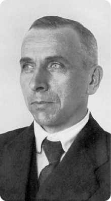 Fotografia em preto e branco do busto de um homem de pele clara, cabelos curtos e olhos claros. Está vestindo camisa, terno e gravata.