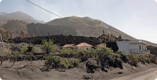 Fotografia de uma paisagem, ao fundo montanhas, à frente, casas, um material escuro cobrindo parte do relevo e chão coberto por cinzas.
