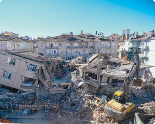 Fotografia de escombros de construções ao redor de prédios e uma escavadeira em meio aos entulhos.