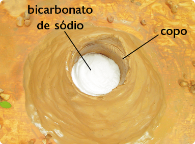 Fotografia de cima do modelo do vulcão, com o copo no centro e um pó branco em seu interior, com a seguinte indicação: bicarbonato de sódio.