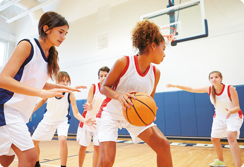 Fotografia de adolescentes de dois times diferentes, jogando basquete, em uma quadra de esporte fechada.