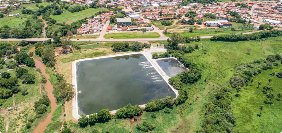 Fotografia aérea de um local aberto com dois tanques de formato retangular, um de tamanho grande, e ao lado, um menor. No entorno há vegetação, um rio, rodovia e casas.