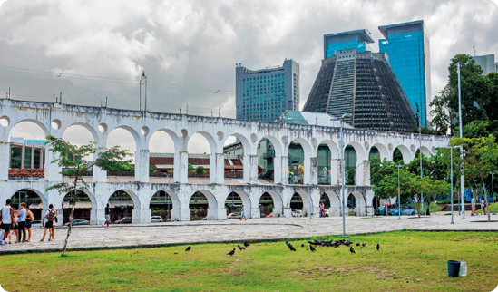 Fotografia de uma construção composta por arcos que são vazados, com dois pavimentos.  No entorno há uma praça com pessoas, gramados e outras construções.