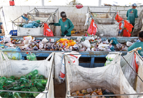 Fotografia de adultos diante de uma estrutura com resíduos recicláveis. Ao redor dessa estrutura, grandes sacos com resíduos recicláveis separados por tipo.