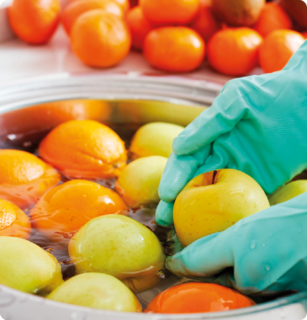 Fotografia de duas mãos vestidas com luvas lavando diversas frutas que estão em um recipiente cheio de líquido incolor.