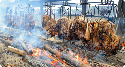 Fotografia de estruturas metálicas apoiadas no chão, com pedaços de carne presos a elas, há alguns centímetros do chão. Próximo à carne, no chão, há pedaços de madeira queimando, com fumaça e fogo visíveis.