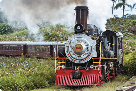 Fotografia de um trem em movimento, à frente, a locomotiva, com uma chaminé de onde sai uma fumaça escura. Um pouco atrás da chaminé também há um local de onde sai uma névoa esbranquiçada. Atrás da locomotiva estão os demais vagões, com janelas.