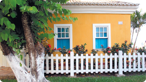 Fotografia de uma casa ao fundo, com duas janelas, uma cerca de madeira e jardim; à frente, uma árvore e gramado.