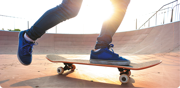 Fotografia dos joelhos para baixo de uma pessoa andando de skate em uma pista de skate. Um pé está em cima do skate e o outro está no ar, para trás, próximo ao solo.