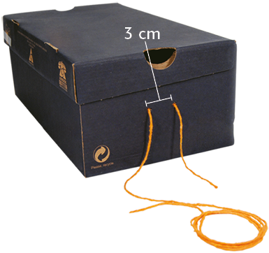 Fotografia. Uma caixa de sapato com dois furos nas laterais menores, atravessados por um barbante. Uma indicação entre os furos indica uma distância de 3 centímetros entre eles.