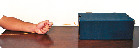 Fotografia. A mão de uma pessoa segurando o barbante e puxando a caixa de sapato horizontalmente em cima de uma mesa.