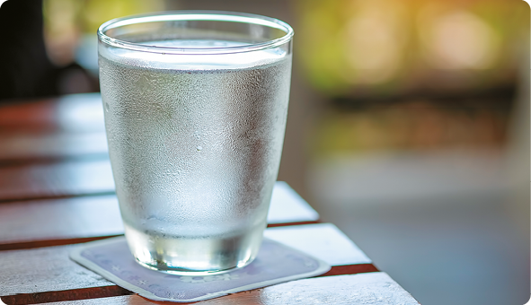 Fotografia. Um copo de água sobre um porta-copos em cima da mesa. O copo apresenta gotículas de água em sua parte externa.