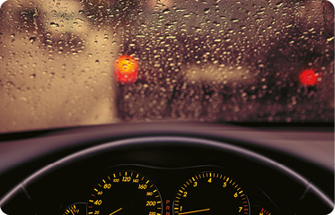 Fotografia. Um para-brisa de carro, visto pelo lado de dentro do veículo, com diversas gotas de água com formato arredondado em sua superfície.