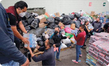 Fotografia. Um local com uma fila de pessoas passando fardos de garrafas de água mineral de mão em mão. As pessoas estão utilizando máscaras cobrindo o nariz e a boca. Entre elas há vários sacos, caixas e fardos de água empilhados em cima de páletes de madeira.