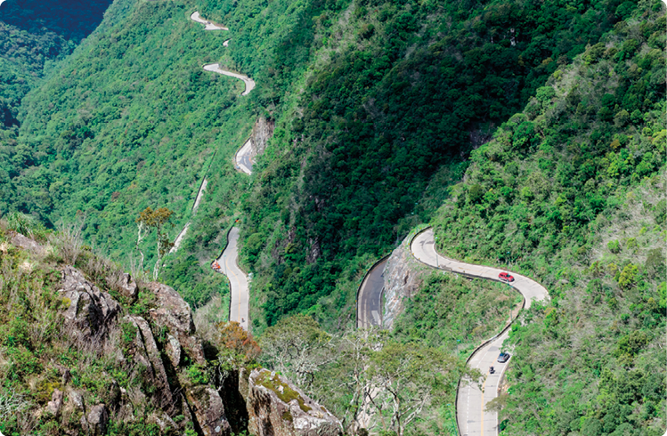 Fotografia. Uma estrada asfaltada que serpenteia por uma montanha íngrime, apresentando curvas acentuadas. A estrada apresenta uma altitude mais elevada em primeiro plano do que ao fundo. Há uma grande quantidade de vegetação ao redor da estrada.