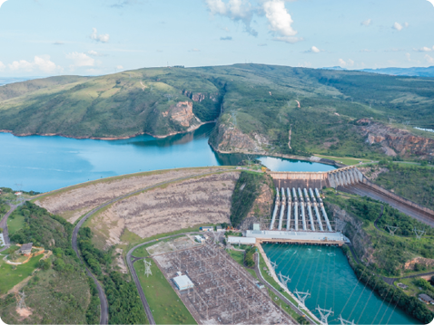 Fotografia. Vista aérea de uma usina hidrelétrica. Na parte inferior direita há um corpo de água e, do lado esquerdo, uma área pavimentada com diversas torres de energia. Acima existe uma parede alta, a barragem, que do lado esquerdo é formada principalmente por rochas, solo e vegetação, e do lado direito, concreto. Acima da barragem há um lago artificial. Na barragem de concreto existem grandes tubulações que partem da parede de concreto e vão até o corpo de água.