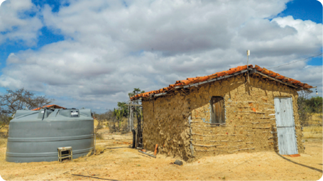 Fotografia. Uma casa de barro, de coloração marrom-claro, com telhado de alvenaria, construída sobre a terra. Ao lado há uma estrutura cilíndrica, de cor cinza. O Sol está iluminando o ambiente, mas há muitas nuvens no céu.