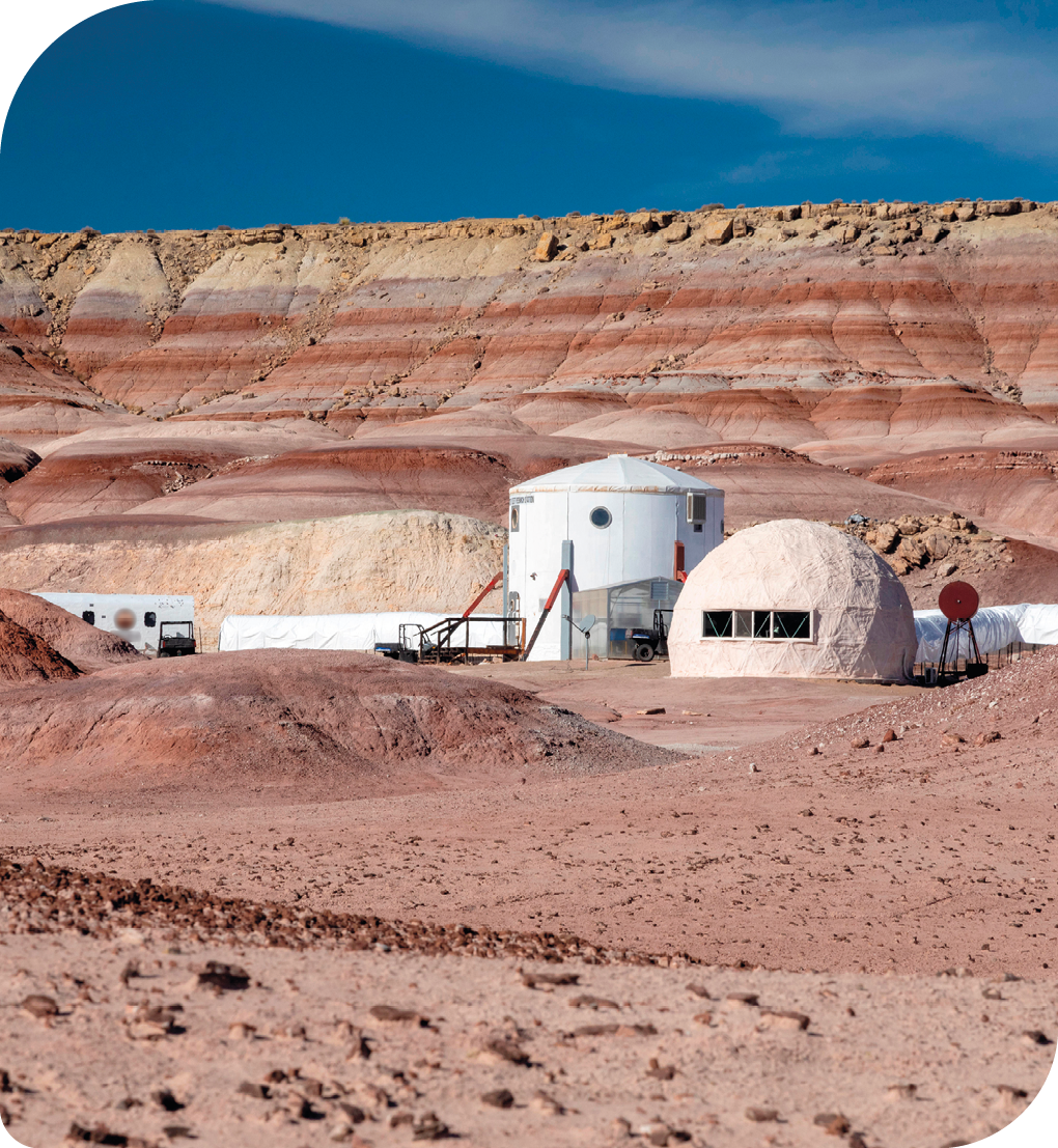 Fotografia. Uma base espacial no meio de um deserto rochoso. Ela está em uma região com terra e pequenos morros, sem vegetação ou corpos de água. No centro, há uma estrutura cilíndrica com janelas e uma porta de frente a uma passarela sustentada por hastes. À direita, há uma estrutura redonda com janelas. À esquerda da estrutura cilíndrica há uma estrutura retangular com janelas. Ao fundo há uma montanha rochosa e acima dela o céu, com poucas nuvens. Entre as estruturas mencionadas há pequenos veículos e estruturas tubulares.