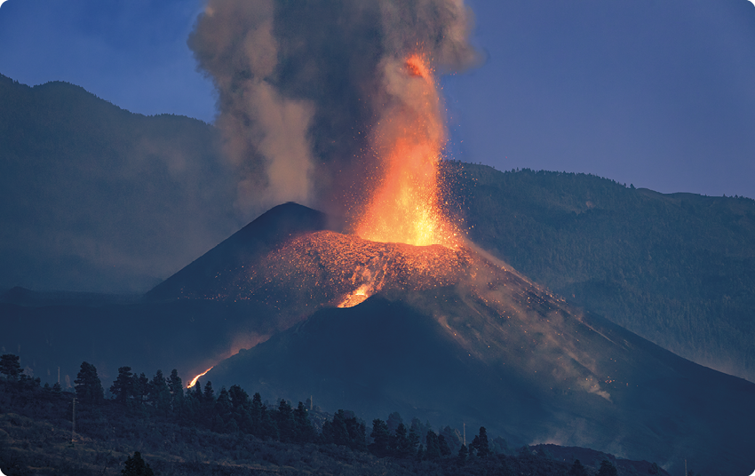 Fotografia de um vulcão com fumaça acima de sua cratera, lava sendo expelida e escorrendo pela sua superfície em formato de cone; ao fundo montanhas, e à frente, árvores.