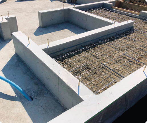 Fotografia da fundação de uma construção, com alicerces de concreto, hastes verticais em suas superfícies e partes do chão concretadas e outras com barras metálicas em formato quadriculado.