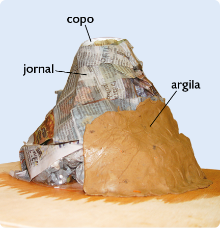 Fotografia de um modelo de vulcão, com formato aproximadamente cônico, feito com folhas de jornal e argila por cima delas. No topo do modelo há um copo encoberto pelas folhas de jornal.
