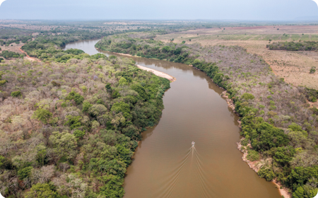 Fotografia. Vista aérea de um rio amarronzado com um barco no meio. Nas laterais, vegetação densa de cor verde e marrom.