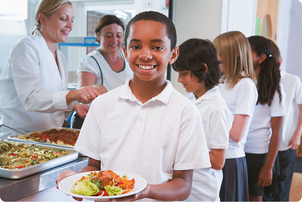 Fotografia de um aluno, à frente, sorrindo e segurando um prato com diferentes tipos de alimentos: saladas e carne. Ao fundo, uma fila de alunos e duas adultas servindo a refeição.