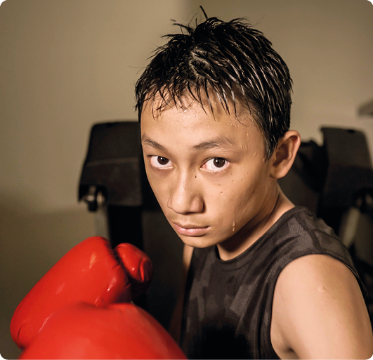 Fotografia de um adolescente com gotas de suor no rosto e cabelo molhado. Ele está usando luvas de boxe, com as mãos em posição de combate e camiseta regata.
