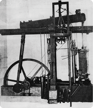 Fotografia de uma máquina composta por um cilindro, à direita, e uma roda, à esquerda, conectados a engrenagens e canos e sustentados por uma estrutura de madeira.