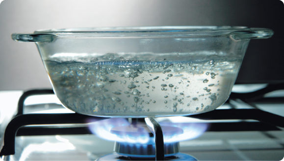 Fotografia de um recipiente de vidro com água sobre a chama de um fogão. Existem diversas bolhas em toda extensão da água.