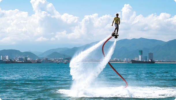 Fotografia. Uma pessoa utilizando um flyboard no meio do mar, há alguns metros acima da água. O equipamento está preso aos pés da pessoa, e dois jatos de água saem da parte inferior em direção à superfície da água. Também há uma mangueira flexível saindo do equipamento e entrando na água do mar.