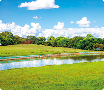 Fotografia. Um parque com muita grama verde e um trecho de água no meio. Ao fundo, há várias árvores. O céu está azul e com várias nuvens.